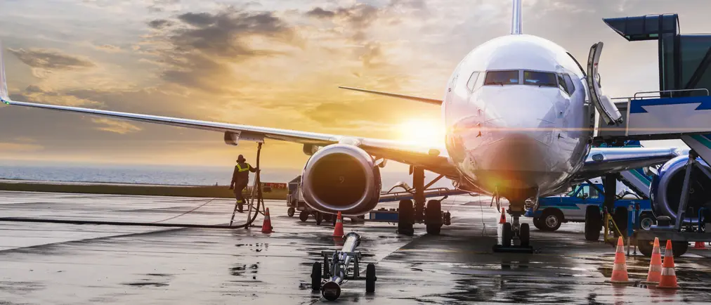 An aeroplane at an airport being refuelled and loaded as the sun sets or rises over the horizon